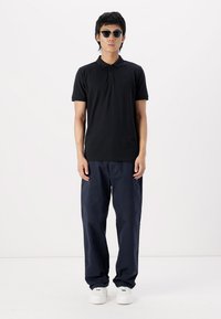 Black polo shirt with short sleeves, paired with loose navy trousers. Model wears round black sunglasses and white sneakers. Minimalist design.