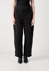 Persona che indossa pantaloni neri a gamba larga con passanti per cintura e tasche anteriori, abbinati a stivali neri con punta a punta sopra una semplice sfondo bianco.