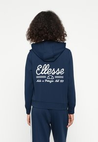 Sweat à capuche bleu marine avec le logo et le texte "Ellesse" blancs au dos, doté d'une capuche avec cordon et de poignets élastiques. Tissu doux.