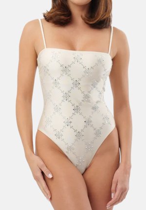 Donna che indossa un costume intero bianco con sottili spalline e motivo floreale diamantato argento, che posa su uno sfondo chiaro e uniforme.