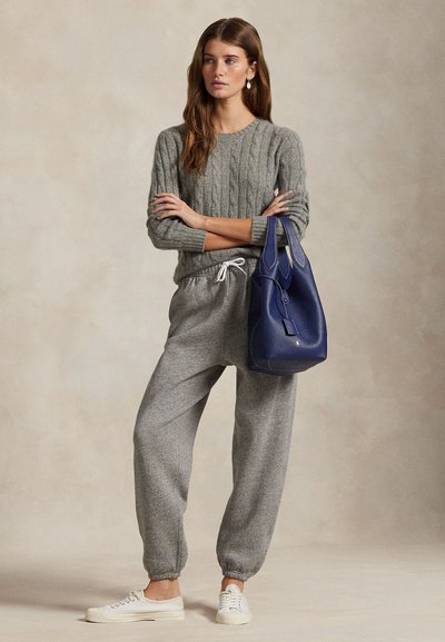 Frau trägt einen grauen Strickpullover mit Zopfmuster und graue Jogginghose, hält eine große blaue Ledertasche und steht mit verschränkten Armen.