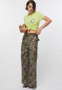 Neonowo zielony krótki t-shirt z grafiką serca, zestawiony z brązowymi moro spodniami typu cargo ozdobionymi zielonymi wzorami liści oraz dużymi bocznymi kieszeniami.