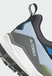 Chaussure avec une tige en maille noire, des accents bleu clair et une semelle intermédiaire grise. Semelle extérieure blanche texturée notable et marquage TERREX bien visible.