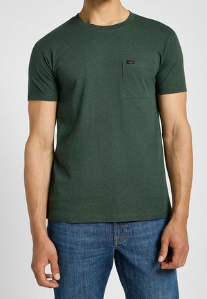 Mężczyzna ubrany w ciemnozieloną koszulkę z krótkim rękawem i kieszonką na piersi oraz niebieskie dżinsy, widoczny od szyi do bioder na jednolitym tle.