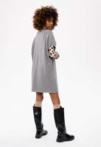 Szara, oversize sukienka z krótkimi rękawami. Wewnętrzne rękawy mają czarno-beżowy wzór w kropki. Modelka nosi sięgnące do kolan czarne botki.