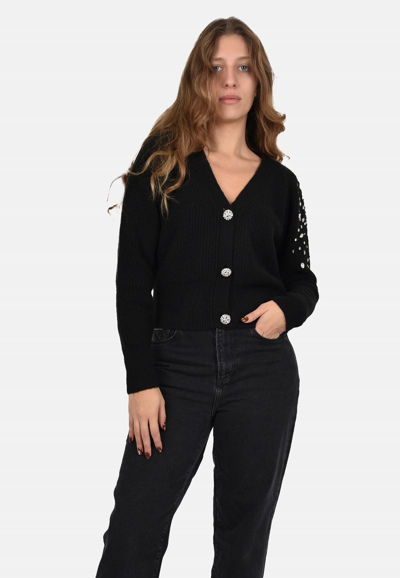 Cardigan nero con una texture a coste, scollatura a V e dettagli decorativi con bottoni. Le maniche presentano delle decorazioni. Indossato sopra jeans scuri.