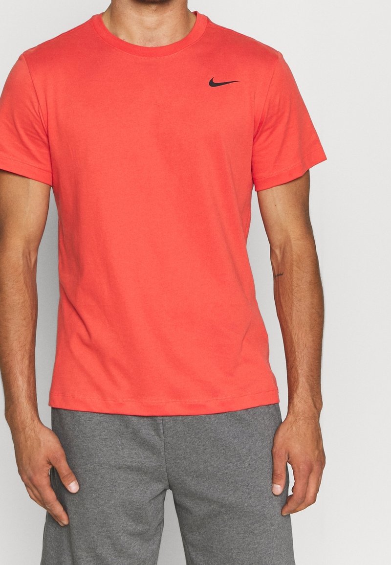 Mężczyzna w jednolitym czerwonym podkoszulku Nike z małym czarnym logo swoosh na piersi oraz szarych szortach sportowych.