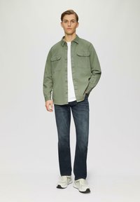 Lichtgroen button-up shirt met twee borstzakken, gedragen over een wit T-shirt, in combinatie met donkerblauwe jeans en lichte sneakers.