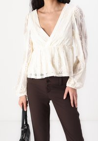 Blusa crema trasparente con pizzo, scollo a V, vita arricciata e maniche lunghe a palloncino. Abbinata a pantaloni aderenti marrone scuro e a una borsa nera.