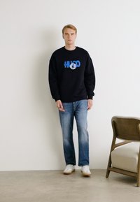 Crni dukserica s plavim logotipom, plave traperice i bijele tenisice. Model stoji uz jednostavan zid; drvena stolica je u blizini.