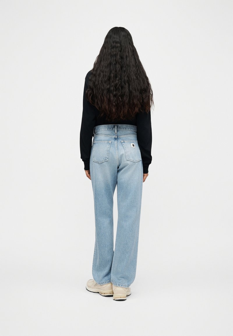 Vrouw met lang krullend haar, gekleed in lichtblauwe jeans met hoge taille, zwart shirt met lange mouwen en beige sneakers, staand met de rug naar de camera op een witte achtergrond.