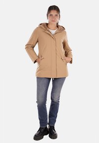 Giacca impermeabile beige con cappuccio, dotata di tasche frontali e taglio dritto. Indossata sopra una maglietta bianca e abbinata a jeans blu e stivali neri.