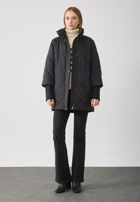 Schwarze gesteppte Parka mit hohem Kragen, Druckknöpfen und weit geschnittenen Ärmeln; getragen über einem cremefarbenen Rollkragenpullover und schwarzen Schlaghosen. Strukturierte Oberfläche.