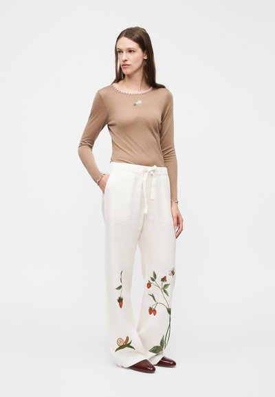 Kvinde iført beige langærmet top og hvide vidde bukser med broderi af jordbær, bi og snegl, stående med hænderne i lommerne.