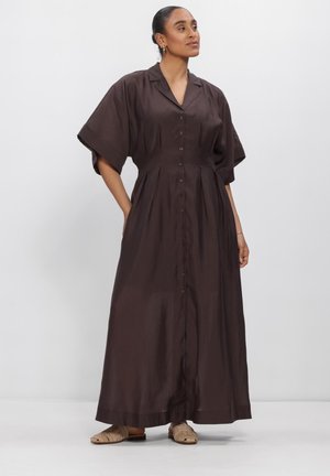 Frau trägt ein dunkelbraunes, knöpfbares Maxikleid mit weiten kurzen Ärmeln und beigen flachen Sandalen, steht vor einem schlichten Hintergrund.