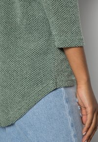 Grau-grüner strukturierter Pullover mit Dreiviertelärmeln, präsentiert über hellblauen Jeans mit ausgefranstem Saum und seitlichen Akzentdetails.