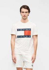 Λευκό βαμβακερό t-shirt με μπλε και κόκκινο λογότυπο "TOMMY JEANS" σε ορθογώνιο σχέδιο. Κοντά μανίκια με στρογγυλή λαιμόκοψη. Casual εφαρμογή.