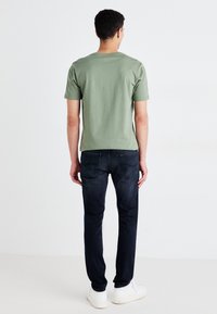 Camiseta de algodón verde con mangas cortas, combinada con jeans de mezclilla azul oscuro y zapatillas blancas, vista desde la parte posterior. Diseño simple.