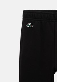 Calças de treino pretas com cintura elástica e bolso lateral, apresentando um pequeno logótipo de crocodilo verde na parte superior da coxa esquerda.