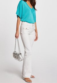 Haut turquoise à manches courtes avec col en V, associé à un pantalon évasé blanc doté de poches avant et de boutons dorés. Sac à main argenté matelassé et sandales.