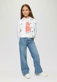 Veste en denim blanc sur un t-shirt graphique avec un motif rouge, assortie à un pantalon large en jeans bleu et des baskets blanches. Fond uni.