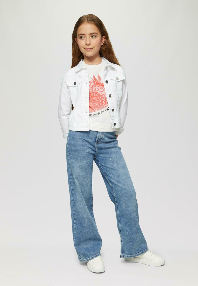 Veste en denim blanc sur un t-shirt graphique avec un motif rouge, assortie à un pantalon large en jeans bleu et des baskets blanches. Fond uni.