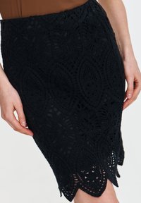 Schwarzer Spitzen-Bleistiftrock mit aufwendigen Blättermustern und einem gewellten Saum. Der Stoff hat eine Struktur mit einem feinen, häkelähnlichen Design.