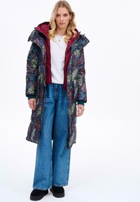 Pohodlný dlouhý kabát s květinovým vzorem v tmavých barvách, s vínovou podšívkou, kapucí a tyrkysovými detaily; v kombinaci s volnými modrými džínami.