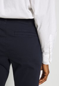 Tummansinisiä puuvillahousuja, joissa on sileä pinta, ja joissa on yksi takatasku. Osittain näkyvissä on valkoinen nappikauluspaita.