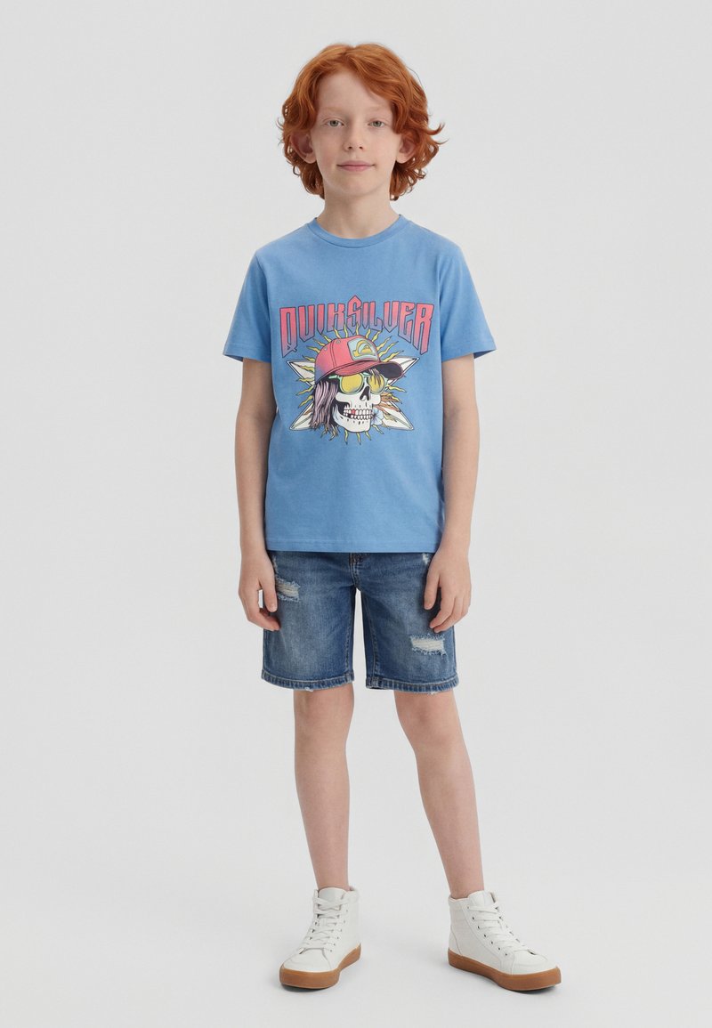 Κοκκινομάλλης αγόρι στέκεται φορώντας μπλε μπλουζάκι με σχέδιο κρανίου και καπέλο, τζιν σορτς και λευκά ψηλά αθλητικά παπούτσια σε απλό φόντο.