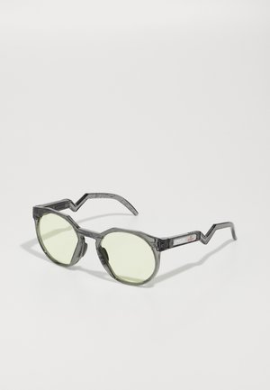 Ochelari de soare rotunzi cu rame negre transparente, lentile verzi și brațe în formă de zigzag. Prezintă sigla Oakley pe braț.