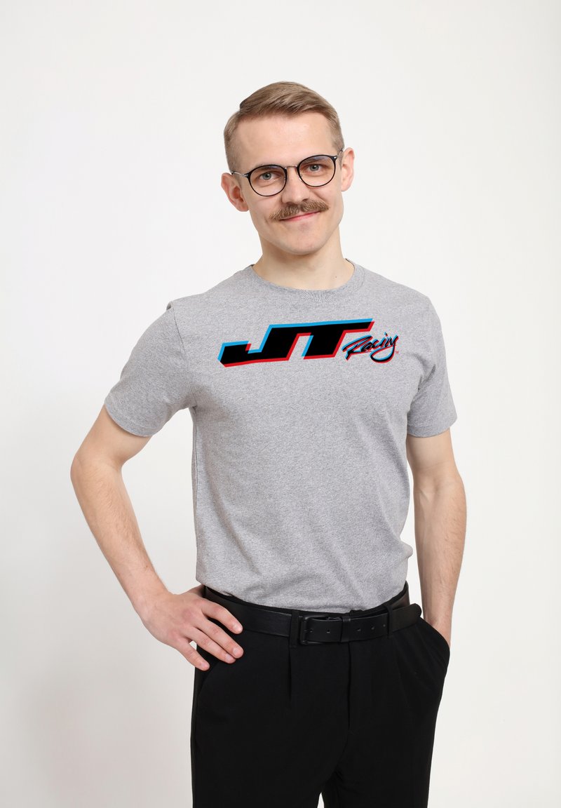 Šedé bavlněné tričko s krátkým rukávem, na kterém je černý a modrý logo "JT Racing" napříč hrudníkem. Standardní střih, nošeno s černými kalhotami.