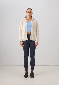 Kreminės spalvos fliso striukė su užtrauktuku, derinama su šviesiai mėlyna trumpa palaidine ir tamsiai mėlynomis tamprėmis. Modelis dėvi juodus sportinius batelius. Paprastas dizainas su kišenėmis.