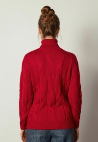 Femme avec une coiffure en chignon tressé portant un pull à col roulé rouge en maille torsadée et un jean bleu, face à un fond clair uni.