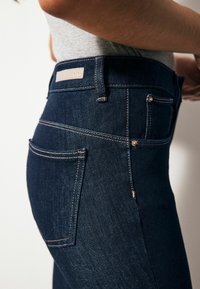 Jeans de tiro alto azul oscuro con costuras doradas, cuatro bolsillos y una etiqueta beige en la cinturilla. Textura de denim suave.