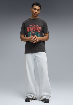 Junger Mann steht mit verschränkten Händen, trägt ein schwarzes "Morocco 2026" Grafik-T-Shirt, weite helle Jeans und schwarze Sneakers.