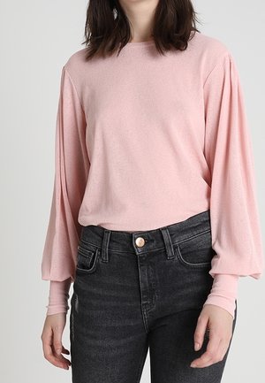 Vrouw die een lichtroze blouse met lange pofmouwen en een zwart hooggetailleerde spijkerbroek draagt tegen een effen achtergrond.