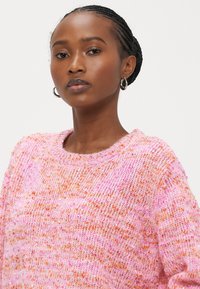 Adīta džemperis rozā un oranžās krāsās ar strukturētu rakstu, apaļu izgriezumu un vaļēju piegriezumu. Smalkas krāsas pūkas piešķir vizuālu interesi.