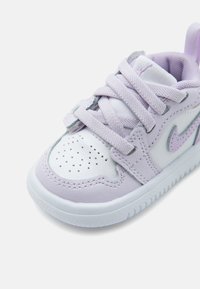 Παιδικό αθλητικό παπούτσι Nike σε λευκό και λιλά, με διάτρητη μύτη, επενδεδυμένο πέλμα και σταυρωτά κορδόνια σε λευκή σόλα.