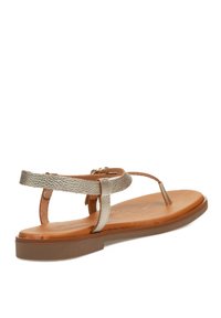 Sandalias doradas y beige con un diseño de tira simple, que presentan una tira delgada en el tobillo y suela plana. Reposan sobre una superficie de madera.