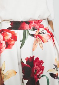 Blommig midikjol med vit bakgrund och röda samt oranga blommor, figursydd midja, plisserad design och slät tygstruktur.