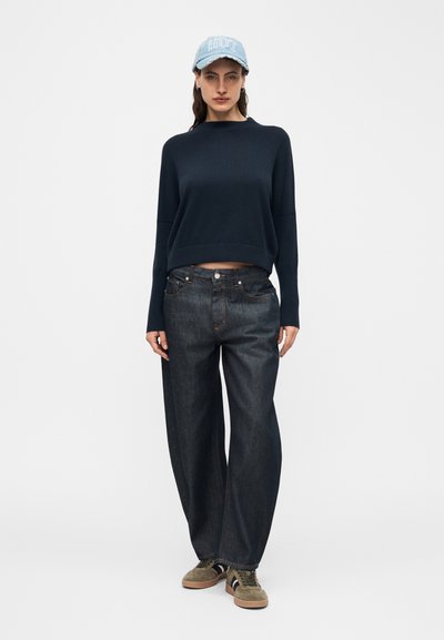 Γυναίκα που φοράει ανοιχτό μπλε καπέλο, φαρδιά μαύρη μπλούζα, παντελόνι με φαρδιά μπατζάκια σκούρου χρώματος και καφέ αθλητικά παπούτσια, στέκεται μπροστά σε απλό λευκό φόντο.