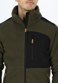 Giacca in pile verde con un pannello in nylon nero sulla spalla, una tasca sul petto con zip e dettagli marroni sui cursori della zip.