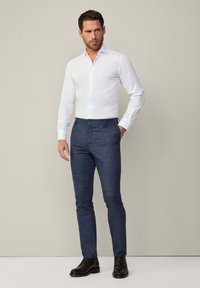 Camicia bianca a maniche lunghe con bottoni abbinata a pantaloni strutturati di colore blu scuro e scarpe in pelle marrone. Collo semplice e design aderente.