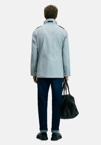 Man wearing light gray jacket, dark blue rolled-up jeans, black shoes, holding black leather bag, standing facing away against white background.