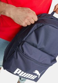 Zaino blu navy con un tessuto testurizzato, caratterizzato da un grande logo Puma bianco e una zip superiore, tenuto da una persona con una maglietta rossa.