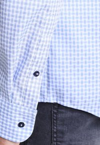 Modrá a bílá kostkovaná látka košile s hladkou texturou, zdobená manžetou s tmavými knoflíky a zaobleným spodním okrajem, doplněná tmavými kalhotami.