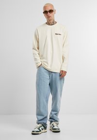 Crème lange mouwen shirt met een bruin logo, lichtblauwe losse spijkerbroek en groen-witte sneakers met veters. Geweven stofdetails zichtbaar.