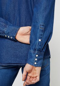 Denim overhemd met lange mouwen, met een gladde textuur, blauwe kleur en drukknopen aan de manchetten. Toont de achterkant en een hand die in de zak rust.