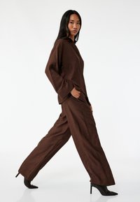 Brun tekstureret outfit bestående af en løs top og bukser, parret med sorte højhælede sko. Enkel design med knapdetaljer.